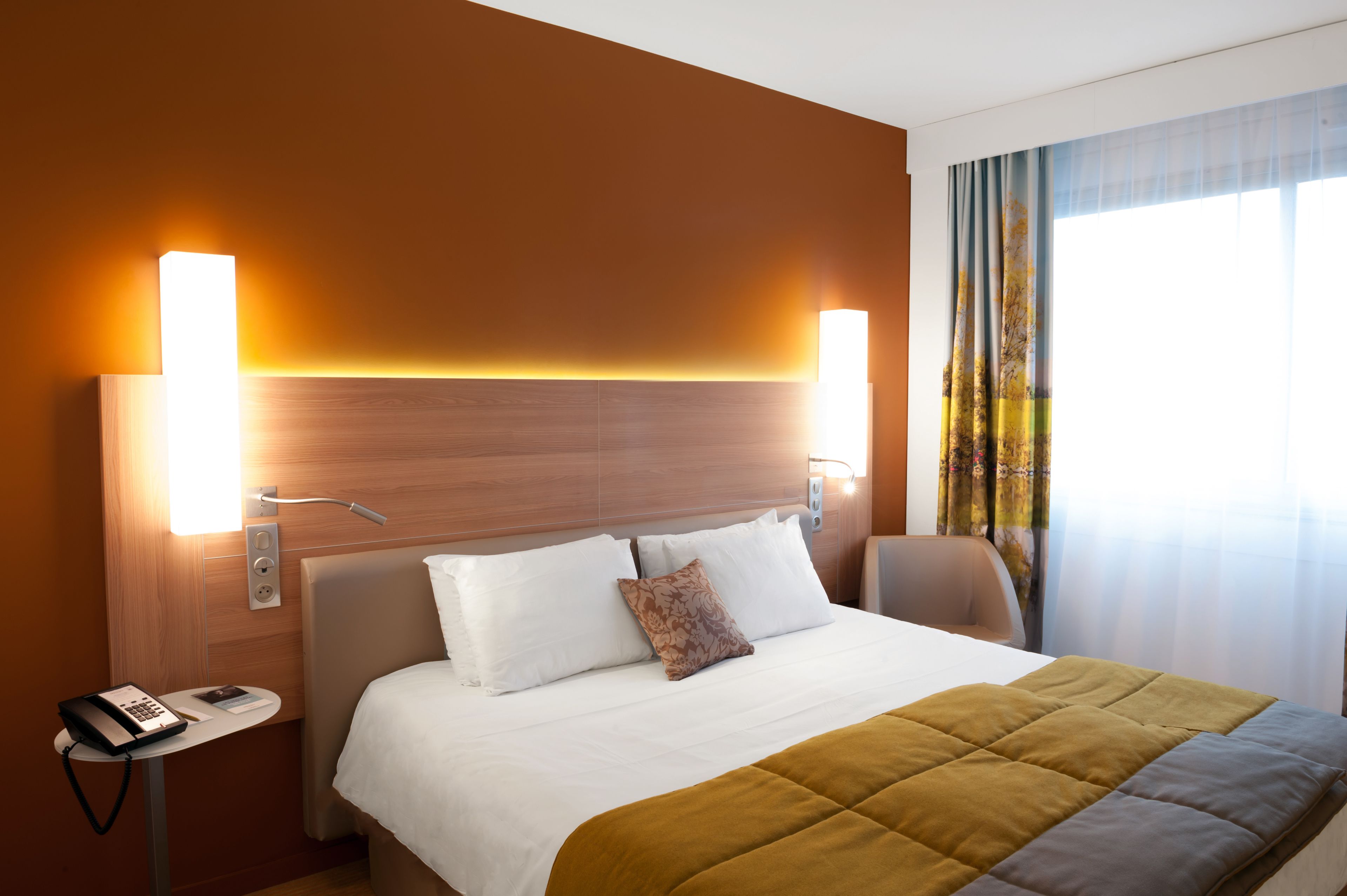 Ein Hotelzimmer mit einem großen Bett, zwei Nachttischlampen, einem kleinen Tisch mit Telefon und einem Fenster mit Vorhängen.