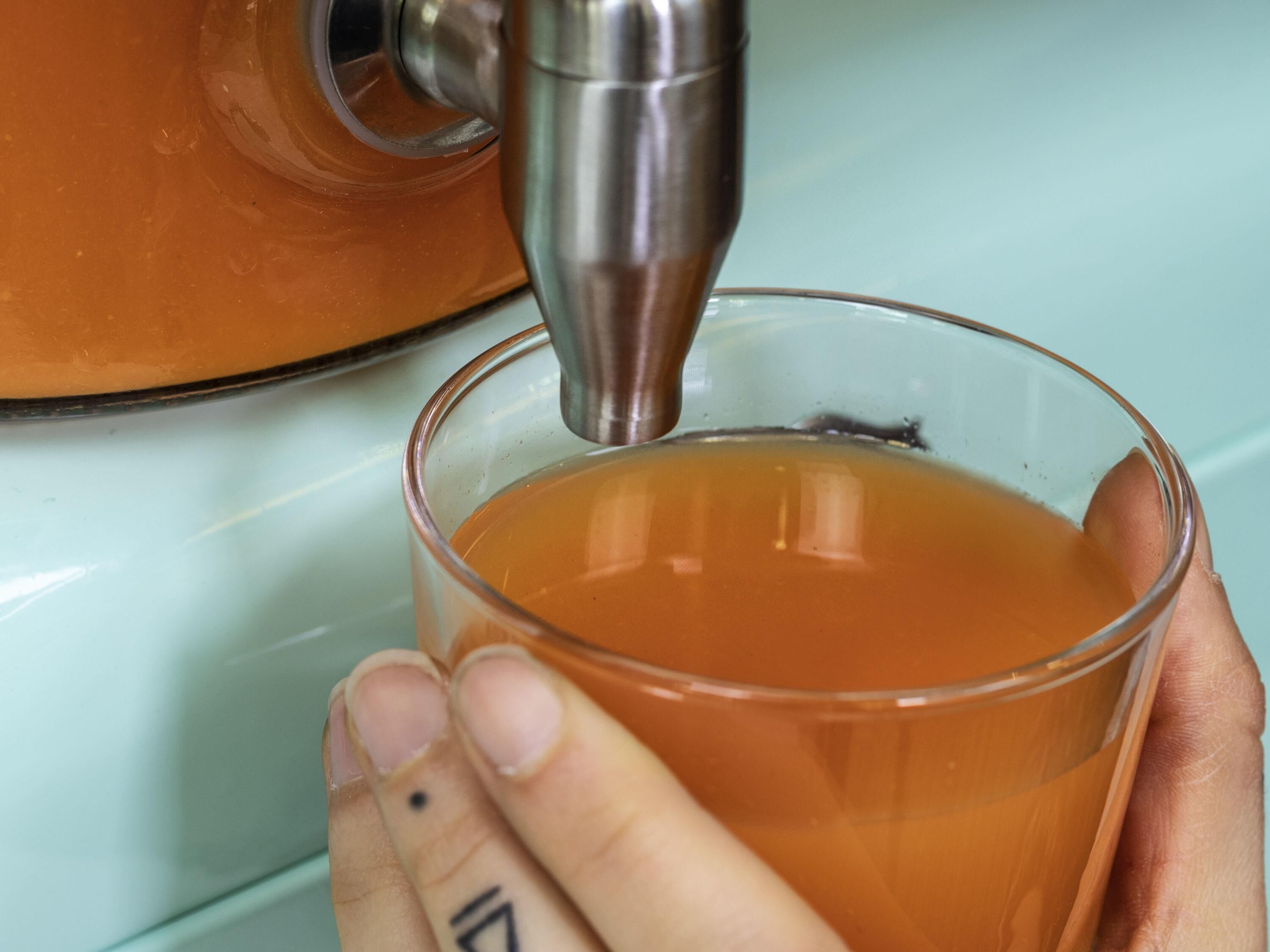 A hand holding a glass with a liquid being poured into it from a metallic container.