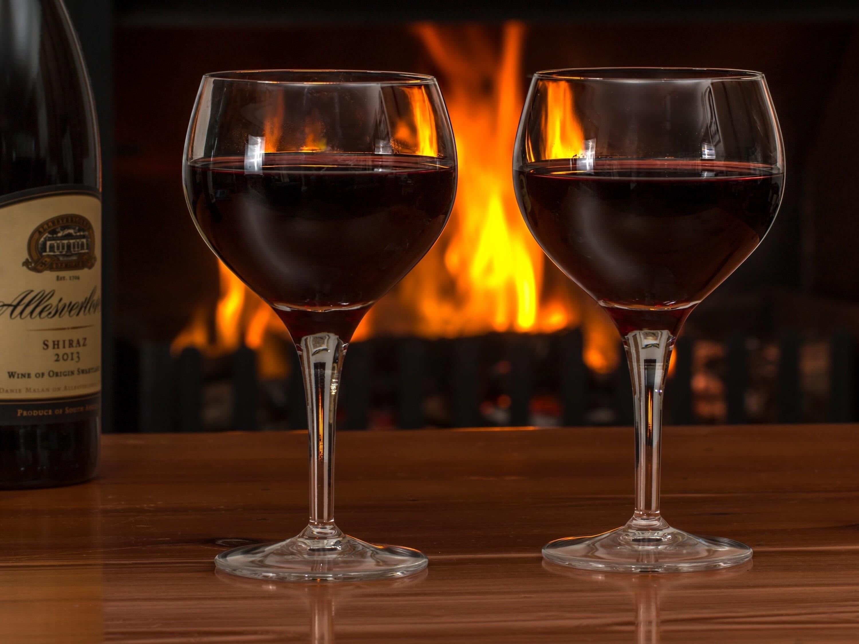 Dos copas de vino sobre una superficie de madera, con una botella de vino y una chimenea al fondo.