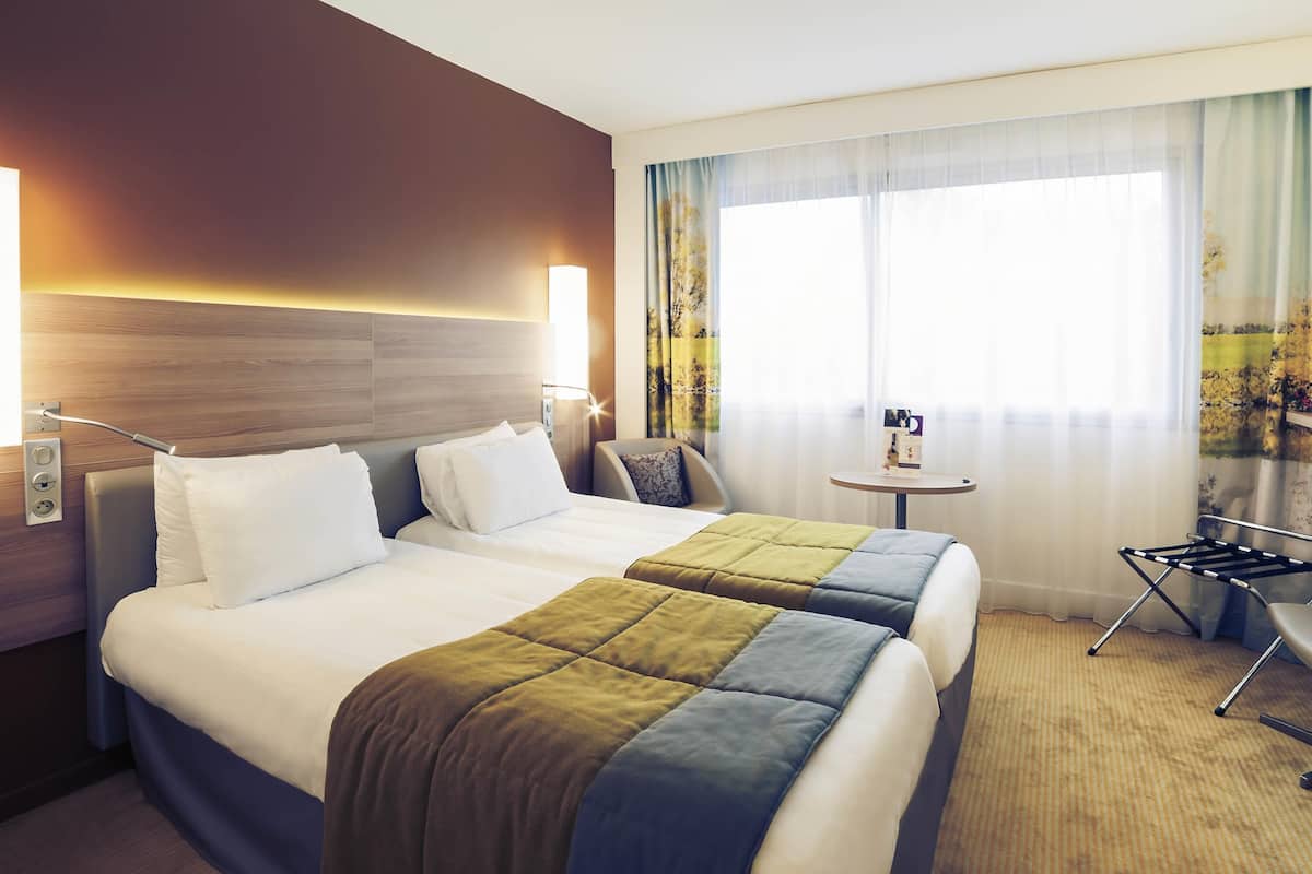 A hotel room with two beds, a small table, a chair, and a window with curtains.