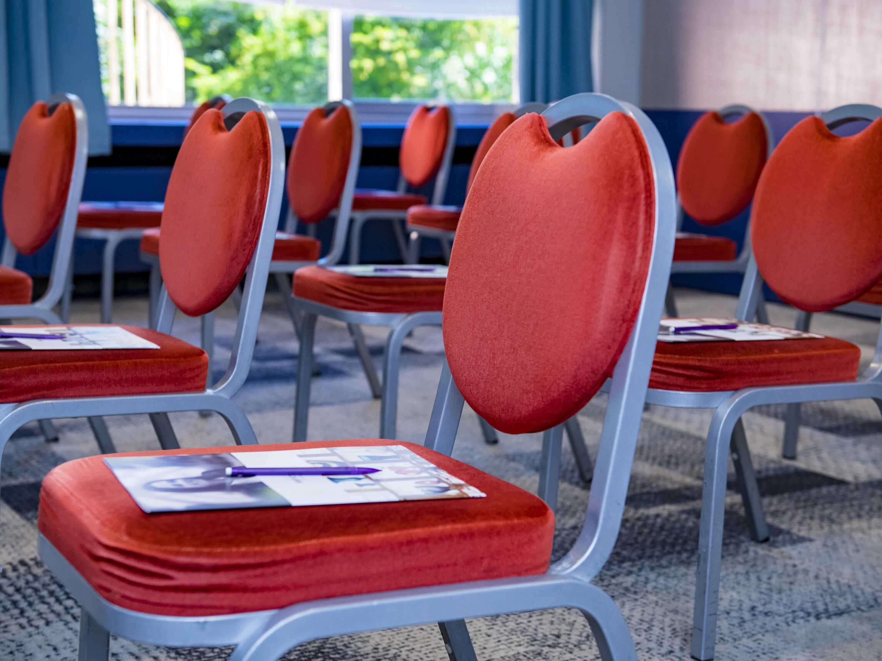 Rows of red cushioned chairs with metal frames, arranged in a spacious room with large windows.