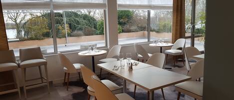 Un restaurante con una decoración moderna, con mesas y sillas blancas, amplios ventanales con vista a zonas verdes y un diseño limpio y minimalista.