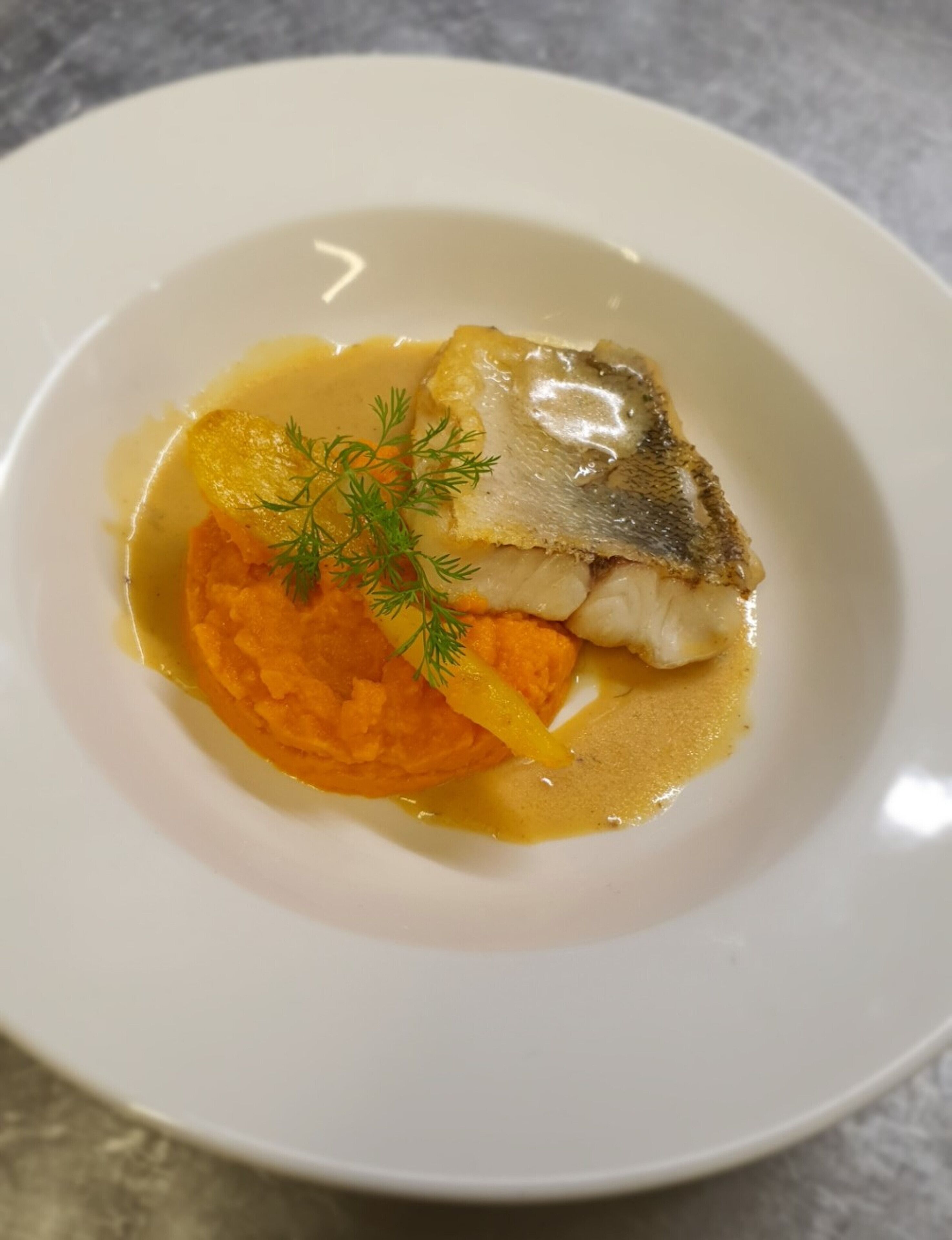 A white plate with a piece of fish, a dollop of orange puree, and a sprig of dill.