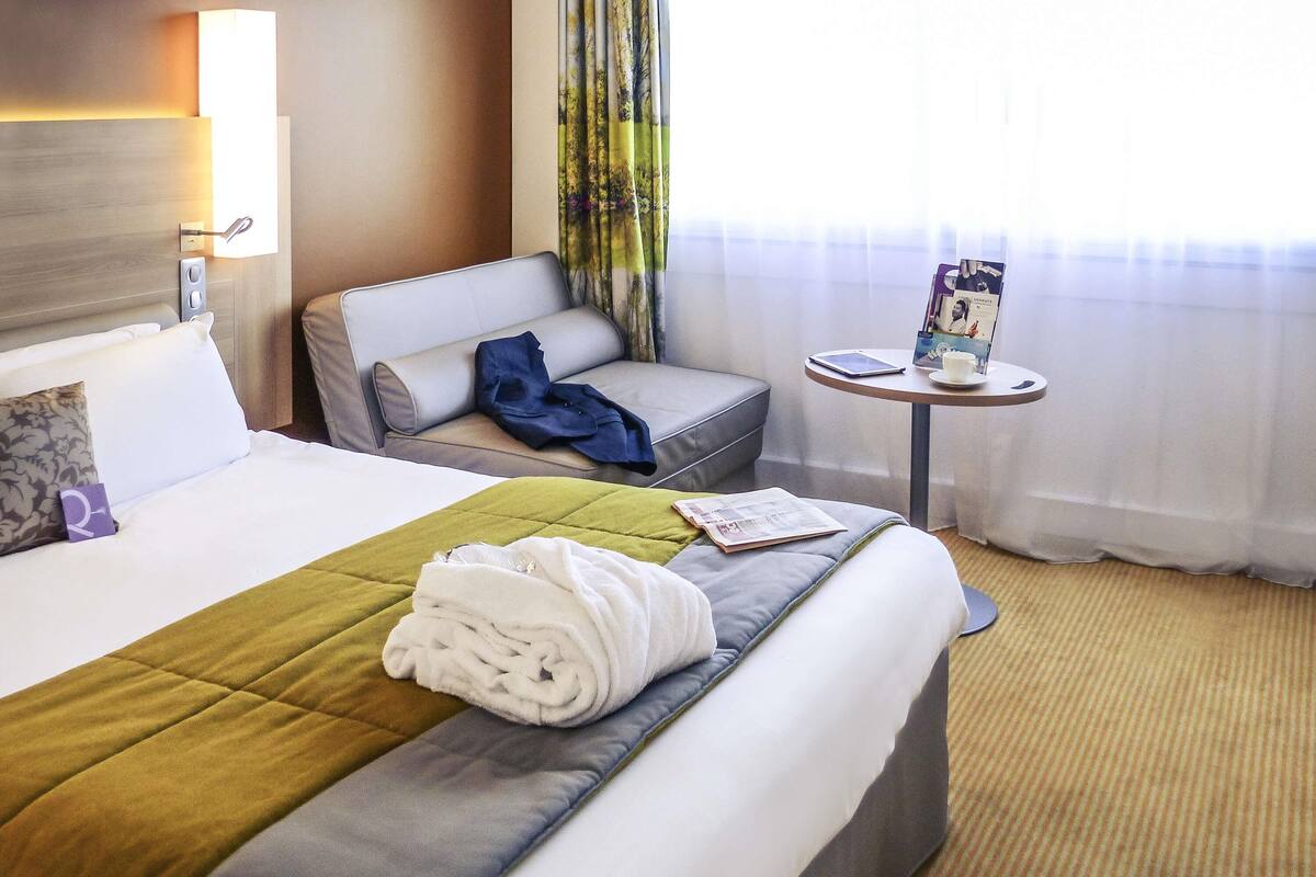 A hotel room with a bed, a sofa, a small table with a magazine, and a window with curtains.