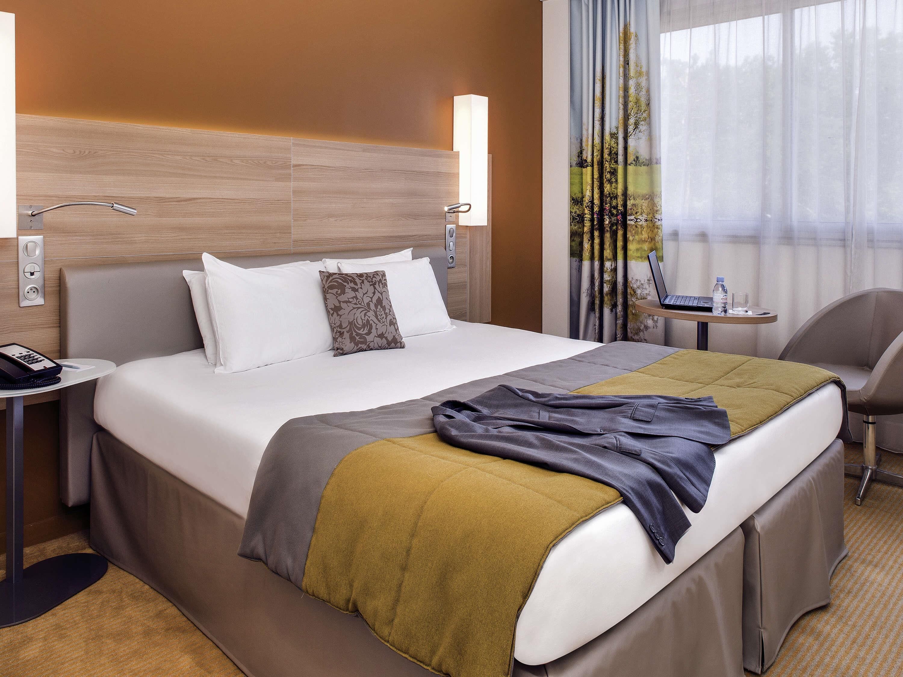 Una camera d'albergo con un letto, una scrivania con un portatile, una sedia e una finestra con tende.