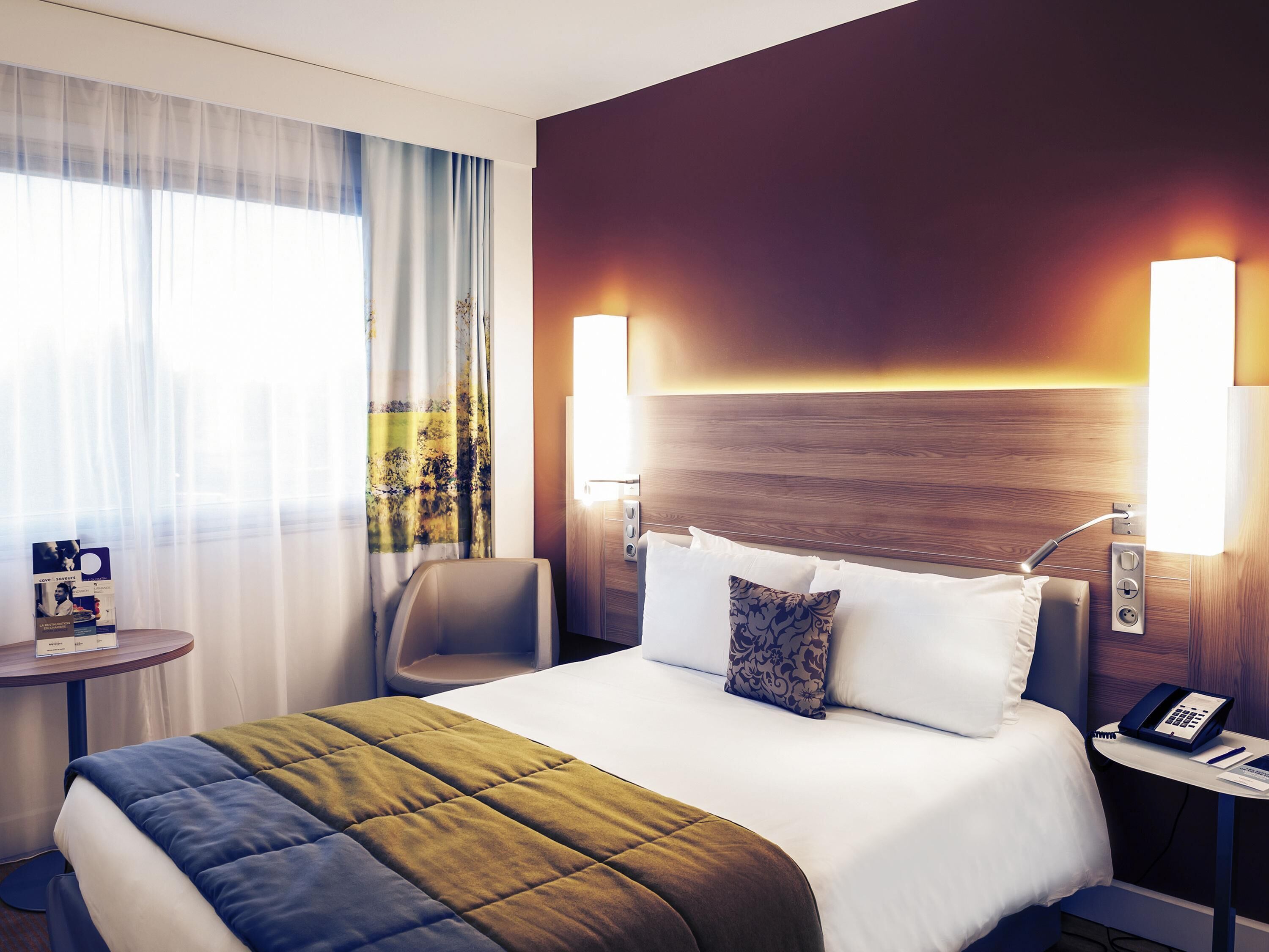 Ein Hotelzimmer mit einem Bett, zwei Nachttischlampen, einem Nachttisch mit Telefon, einem Sessel und einem Tisch mit einer Zeitschrift.