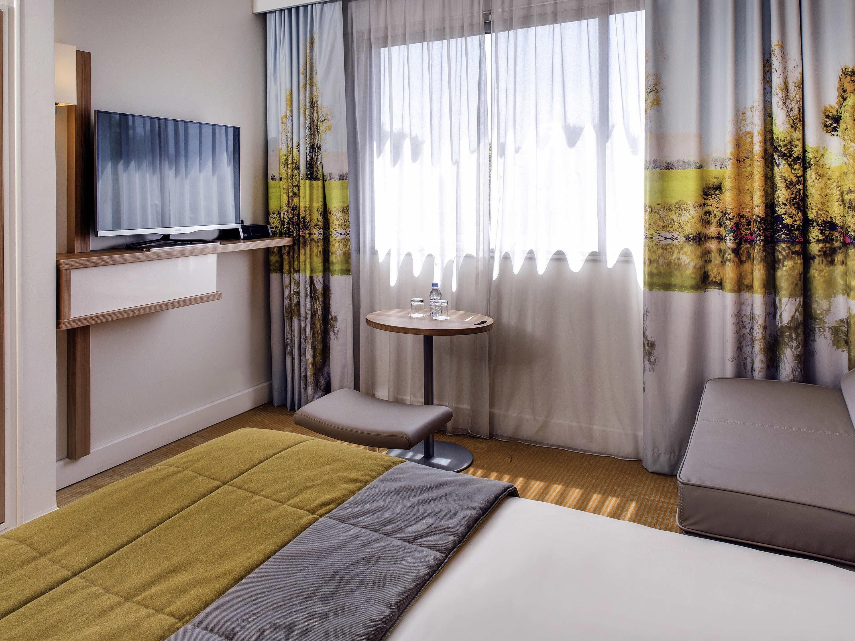 Une chambre d’hôtel avec un lit, une télévision, une petite table avec un lavabo et une fenêtre avec des rideaux légers.
