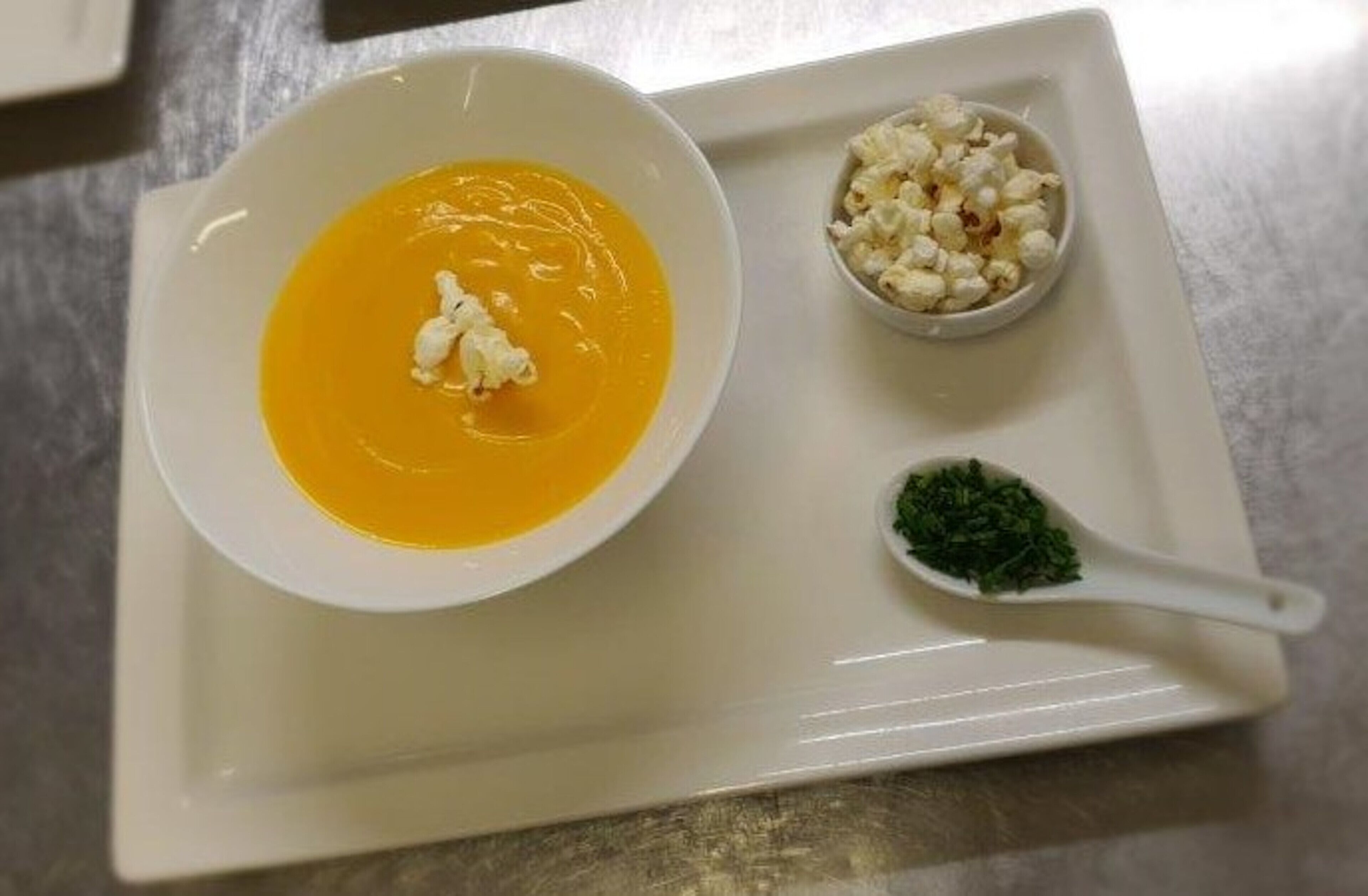 Un plato blanco con un tazón de sopa, un cuenco pequeño de palomitas y una cuchara con guarnición verde.