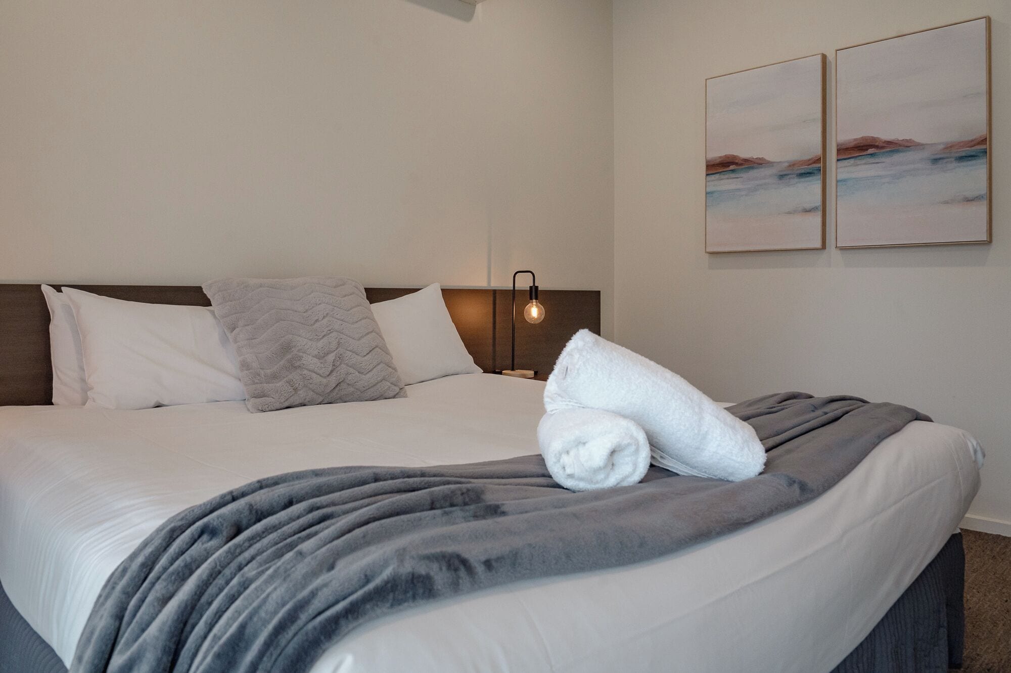 Una cama bien hecha con un edredón gris y almohadas blancas, una lámpara de noche y dos cuadros enmarcados en la pared.
