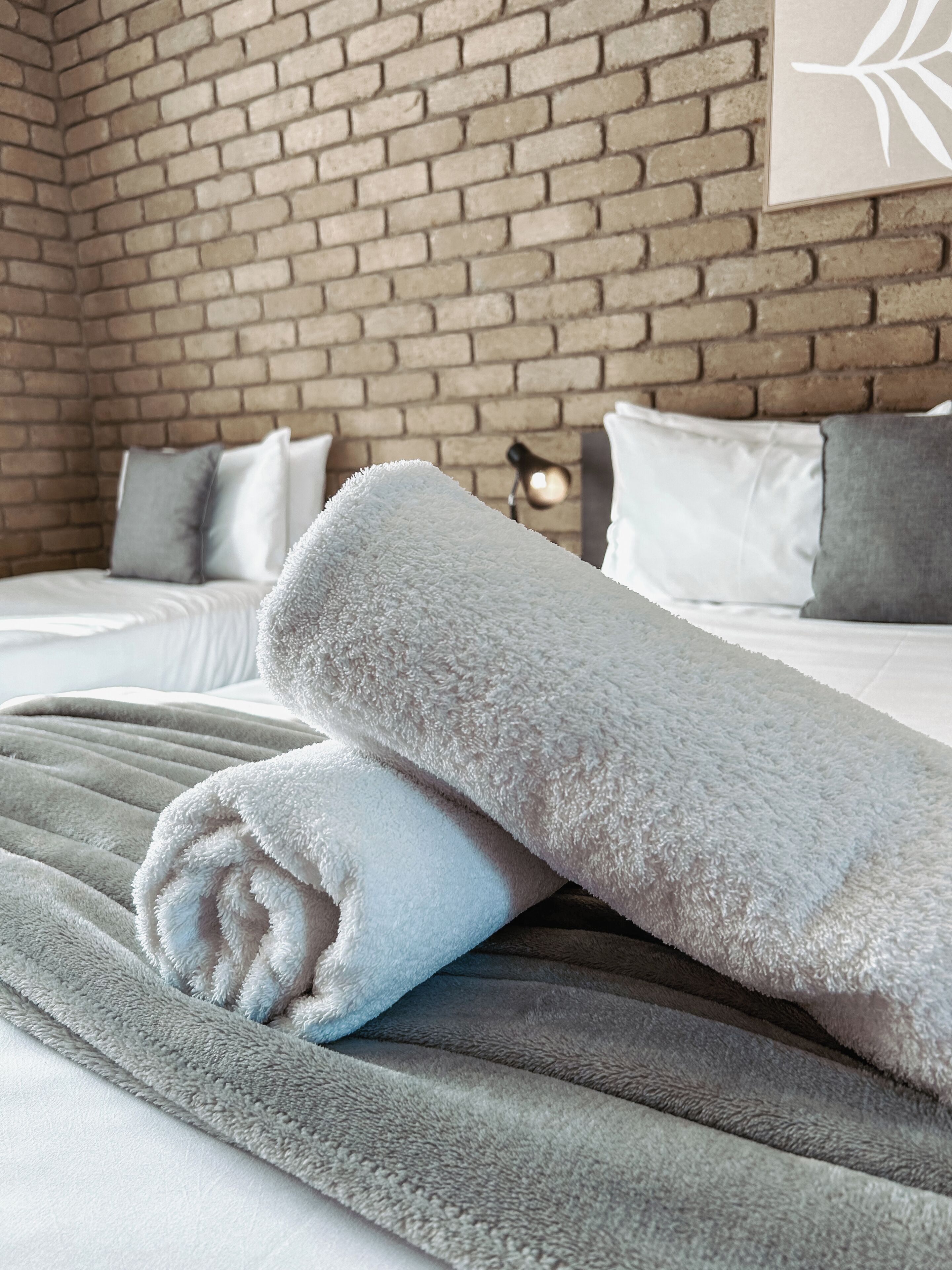 Cama bem arrumada com uma toalha branca enrolada, um cobertor cinza e um muro de tijolos ao fundo.