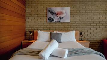 A neatly made bed with white linens and a grey throw blanket, two bedside lamps, and a floral artwork on the brick wall above the bed.