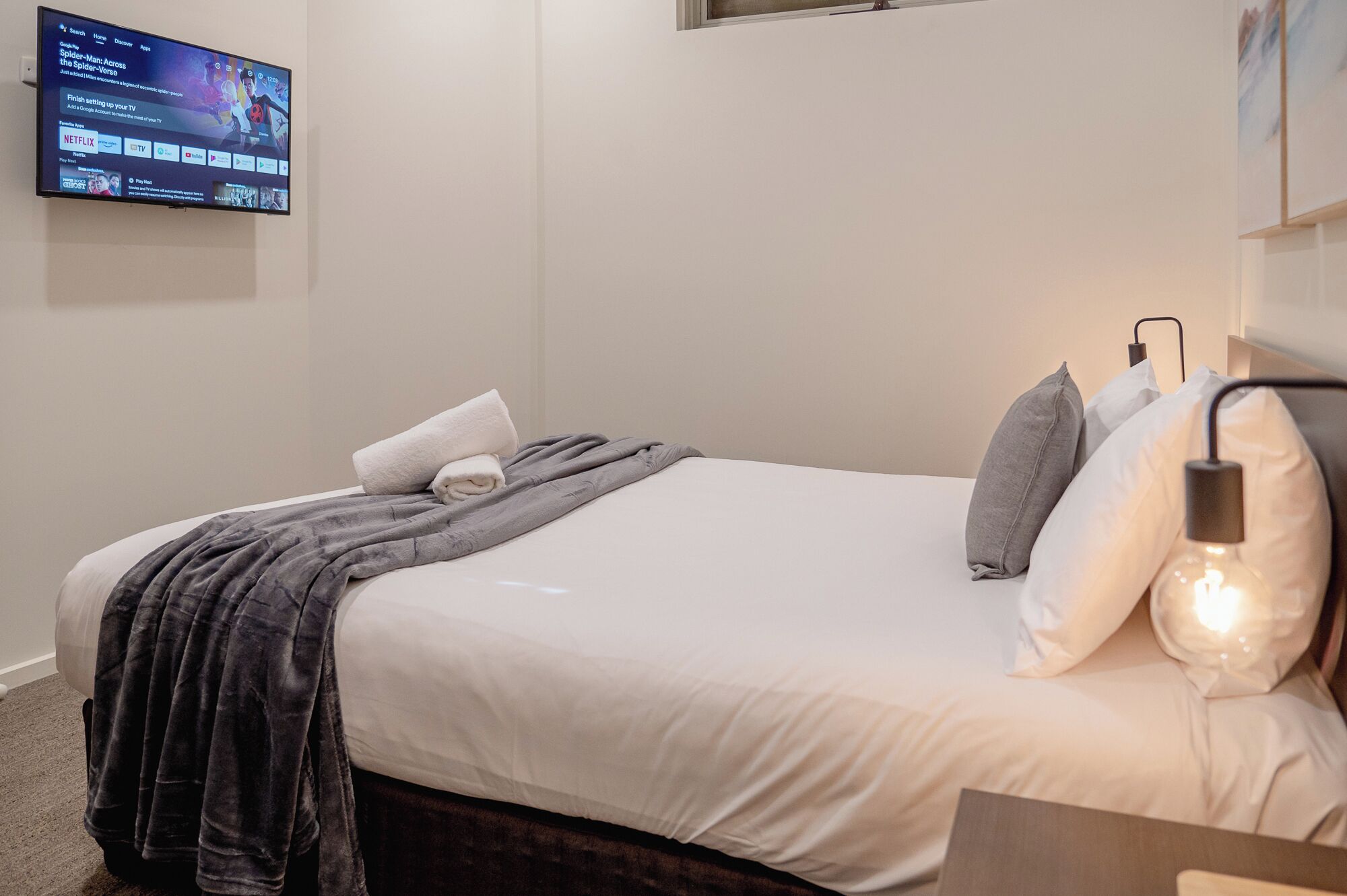 A neatly made bed with a white comforter and a gray blanket, a wall-mounted TV displaying a streaming service interface, and a bedside lamp with a modern design.