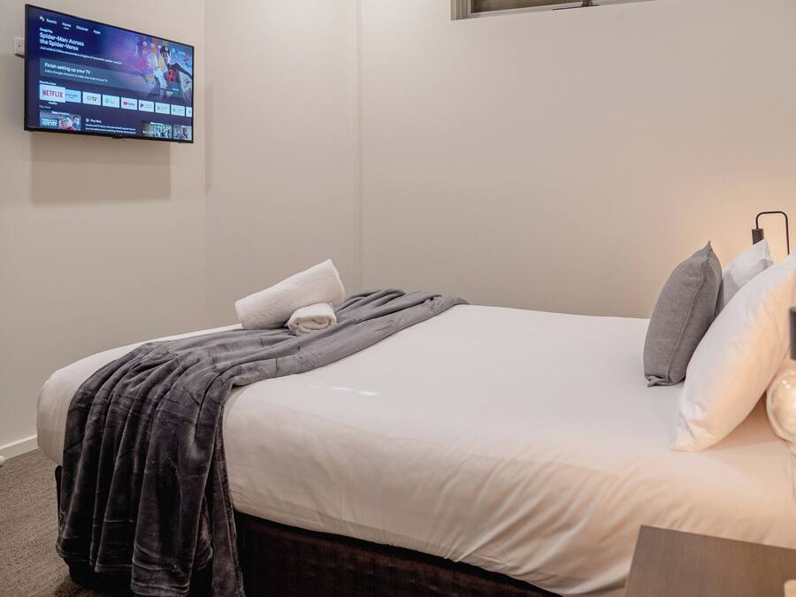 A neatly made bed with a white comforter and a gray blanket, a wall-mounted TV displaying a streaming service interface, and a bedside lamp with a modern design.