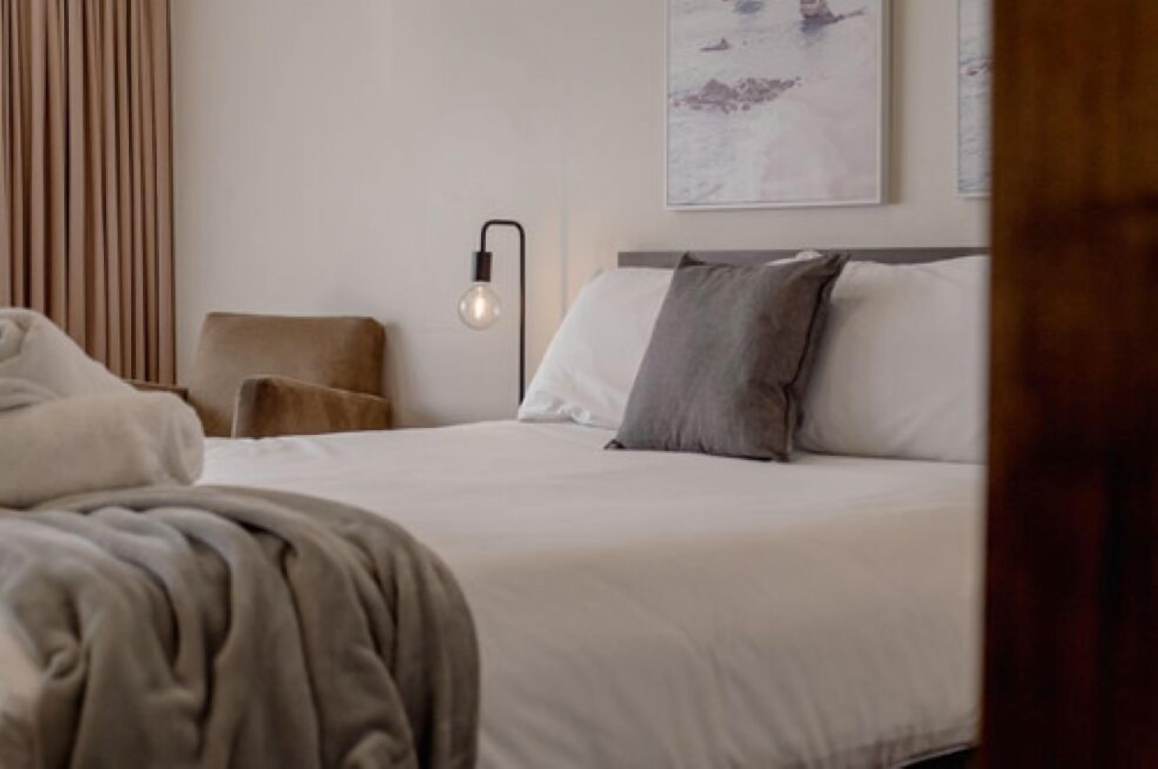Una cama bien tendida con sábanas blancas y un cojín gris, una lámpara moderna fijada a la pared, y un cuadro en la pared.