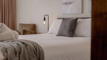 Una cama bien tendida con sábanas blancas y un cojín gris, una lámpara moderna fijada a la pared, y un cuadro en la pared.