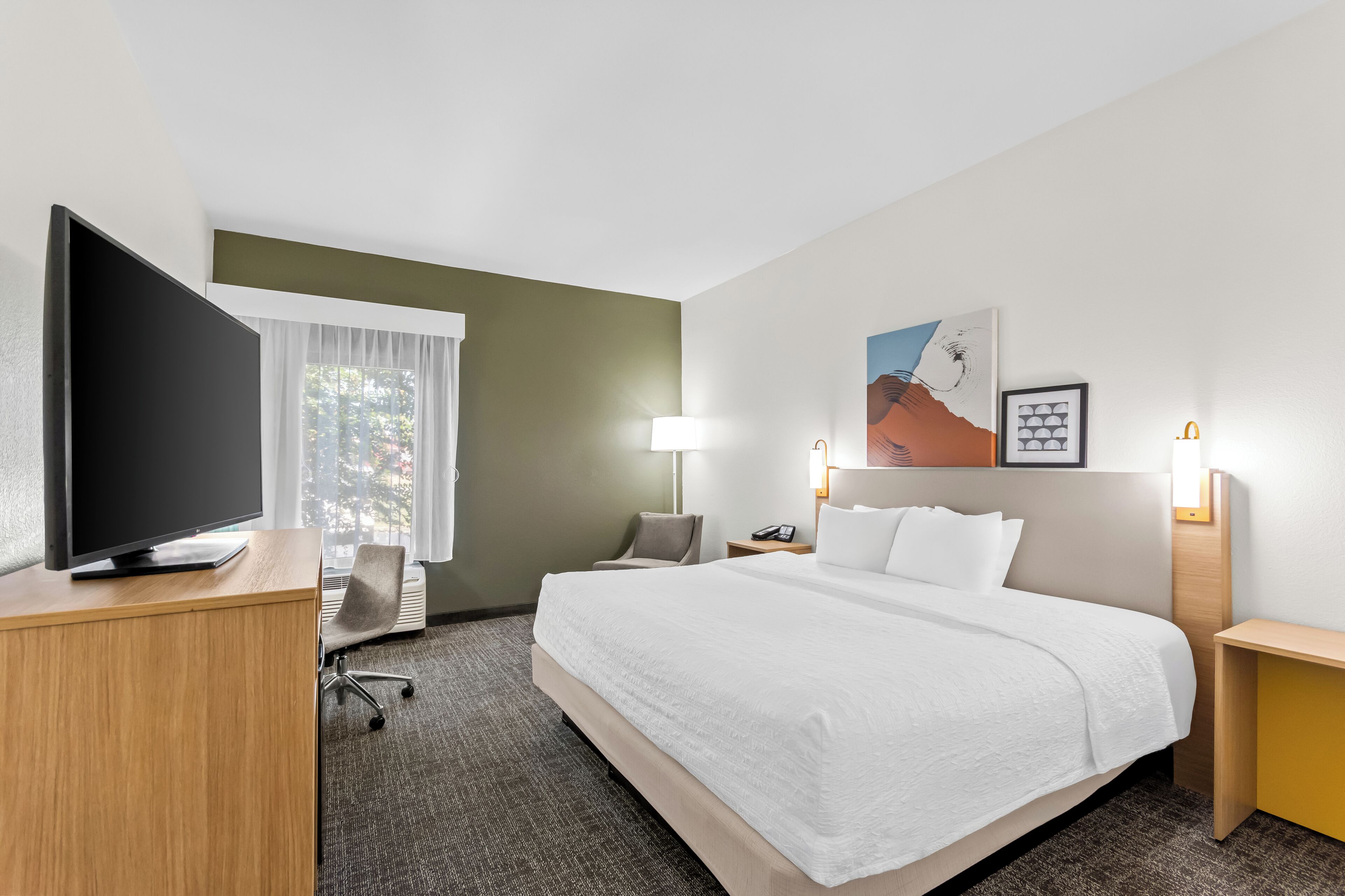 Ein Hotelzimmer mit einem großen Bett, einem Schreibtisch mit Flachbildfernseher, einem Sessel, einer Schreibtischlampe und einem Nachttisch mit Lampe.