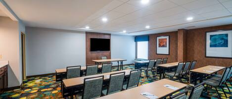 A conference room with multiple tables and chairs, a flat-screen TV mounted on the wall, and a colorful carpet.