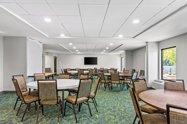 A conference room with multiple tables and chairs, a flat-screen TV on the wall, and a large window providing natural light. - Spark by Hilton Bardstown (Bardstown)