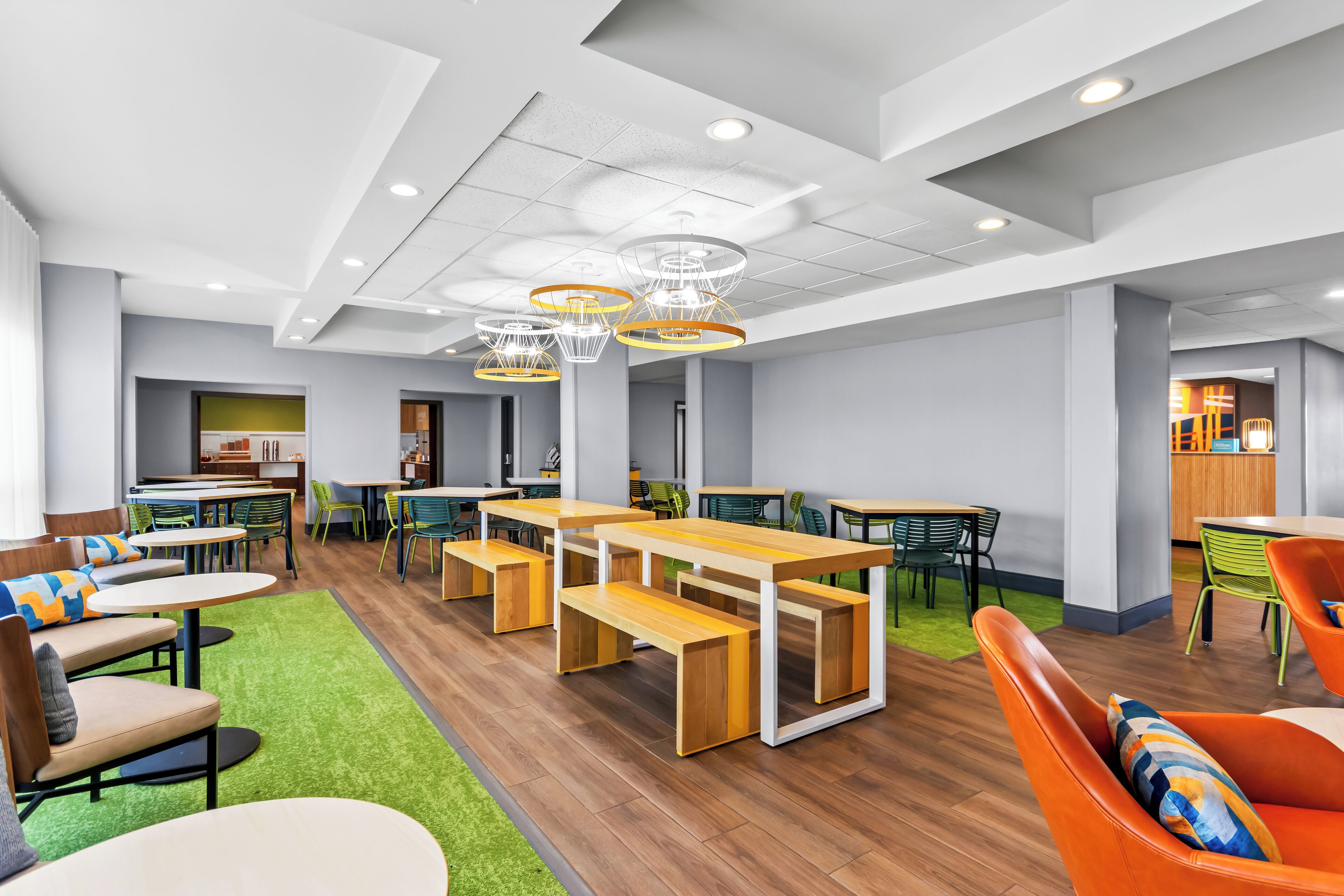 A modern dining area with wooden tables and benches, green chairs, and a bright ceiling with circular light fixtures.