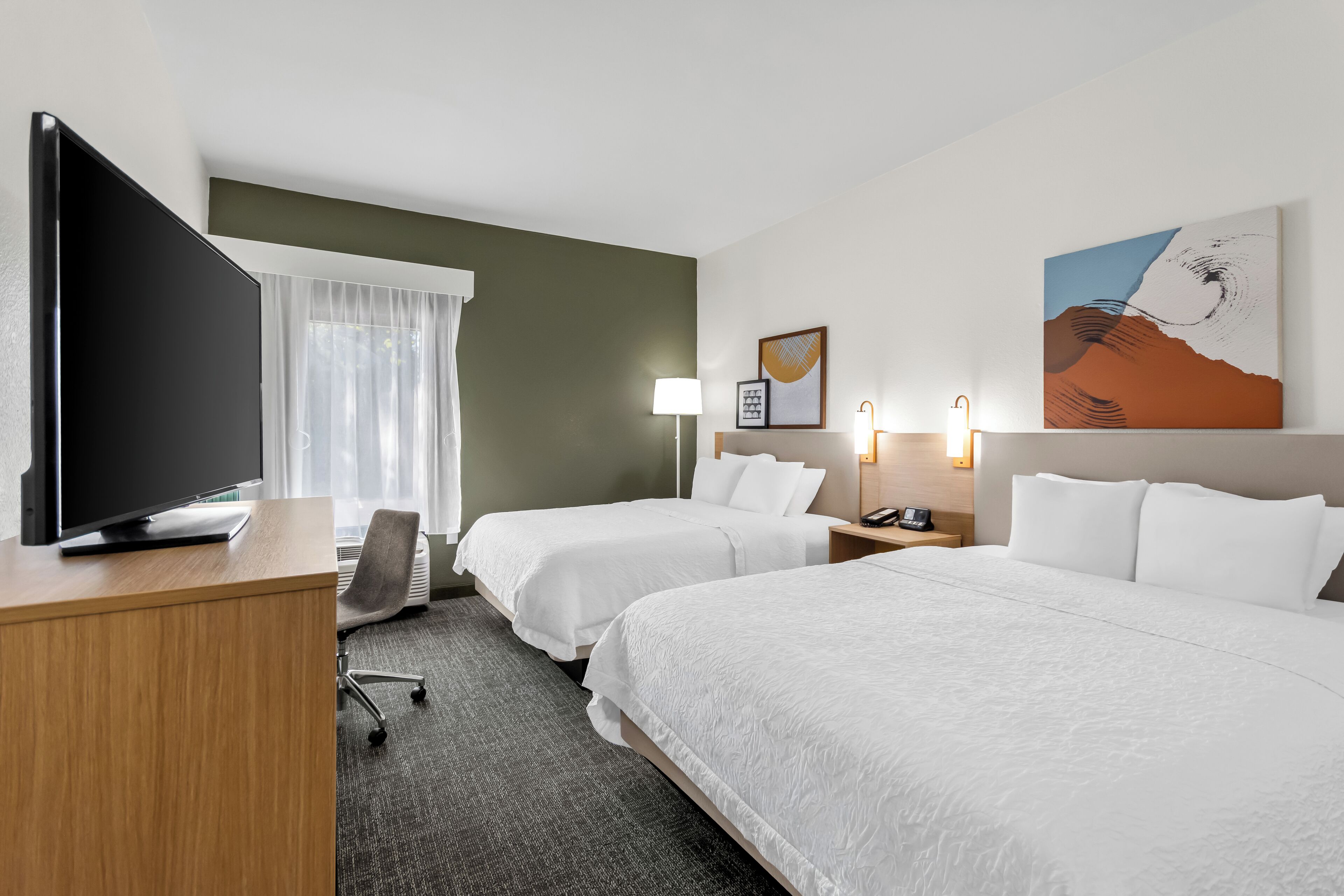 Habitación de hotel con dos camas, un escritorio, un televisor, una lámpara y un cuadro en la pared.