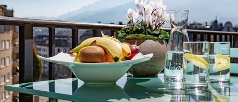 A table with a bowl of fruit, a vase with flowers, and glasses of water on a balcony with a mountain view.