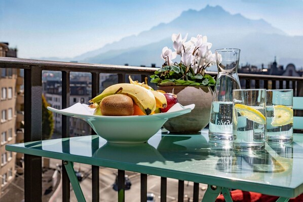 A table with a bowl of fruit, a vase with flowers, and glasses of water on a balcony with a mountain view.