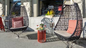 Deux fauteuils à bascule en osier noir avec des coussins roses, une petite table rouge ornée d’un pot de fleurs et d’un verre de vin rouge, et une enseigne de restaurant en arrière-plan.
