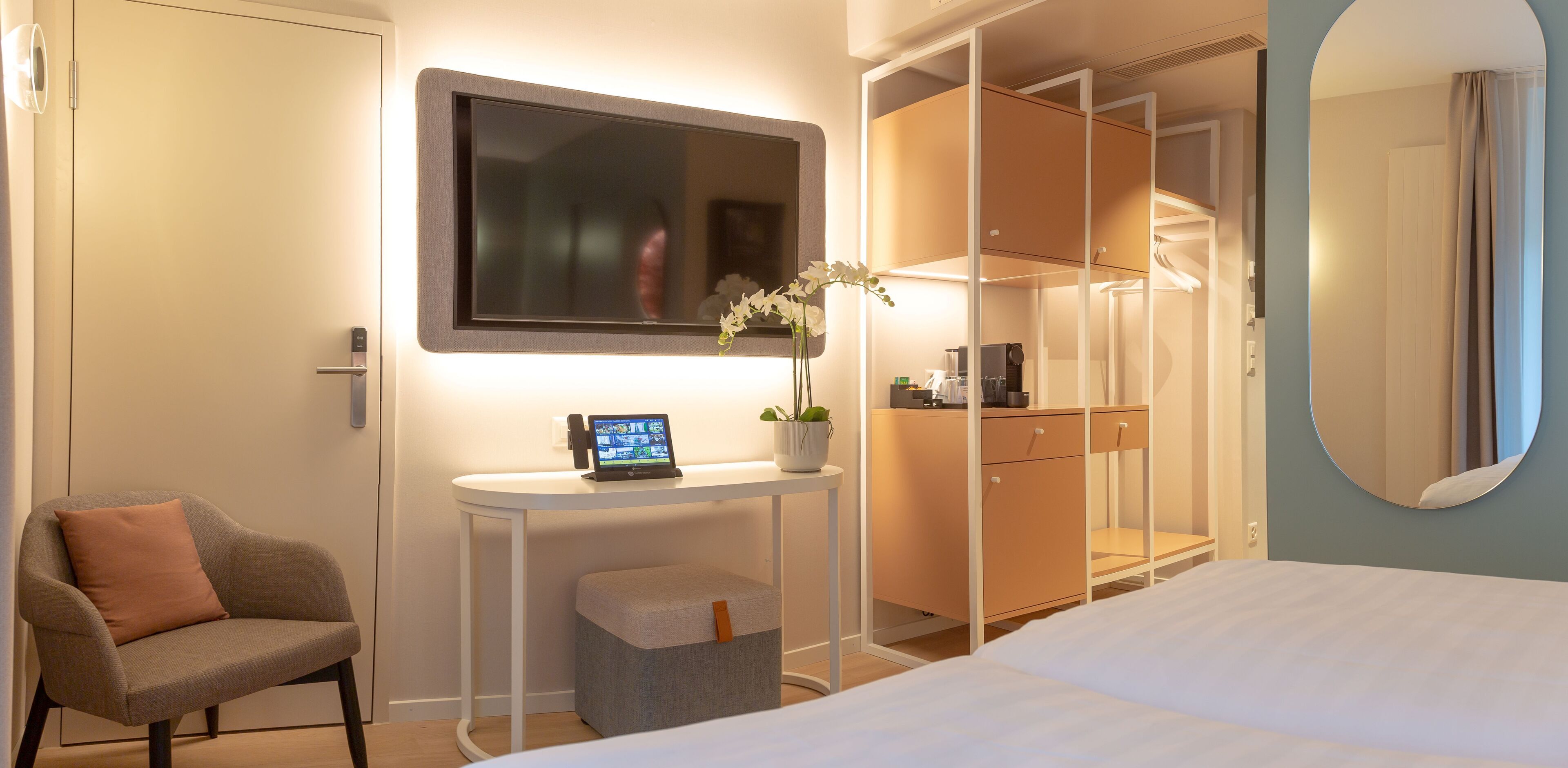 A modern hotel room with a bed, a desk with a laptop, a chair, a wardrobe, and a mirror.