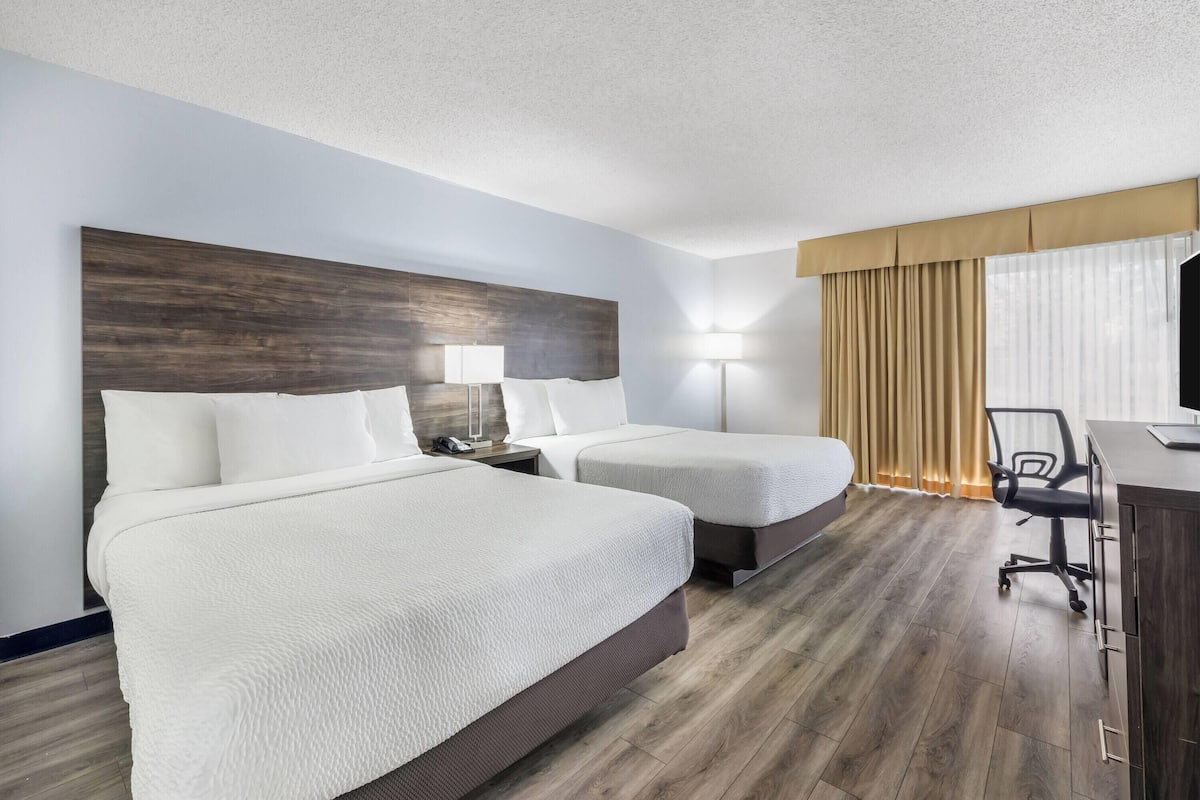 A hotel room with two beds, a desk with a chair, a lamp, and a large window with curtains.