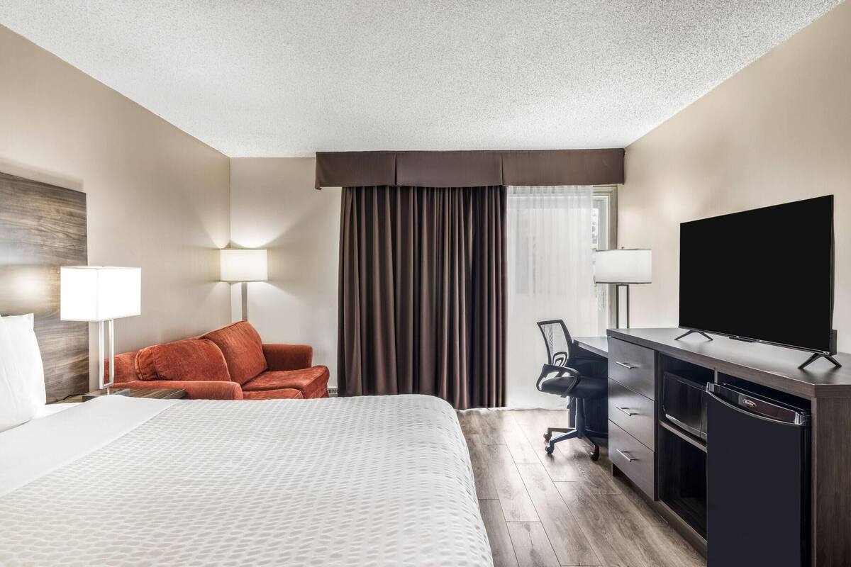 A hotel room with a bed, a desk with a television, a chair, and a desk lamp.