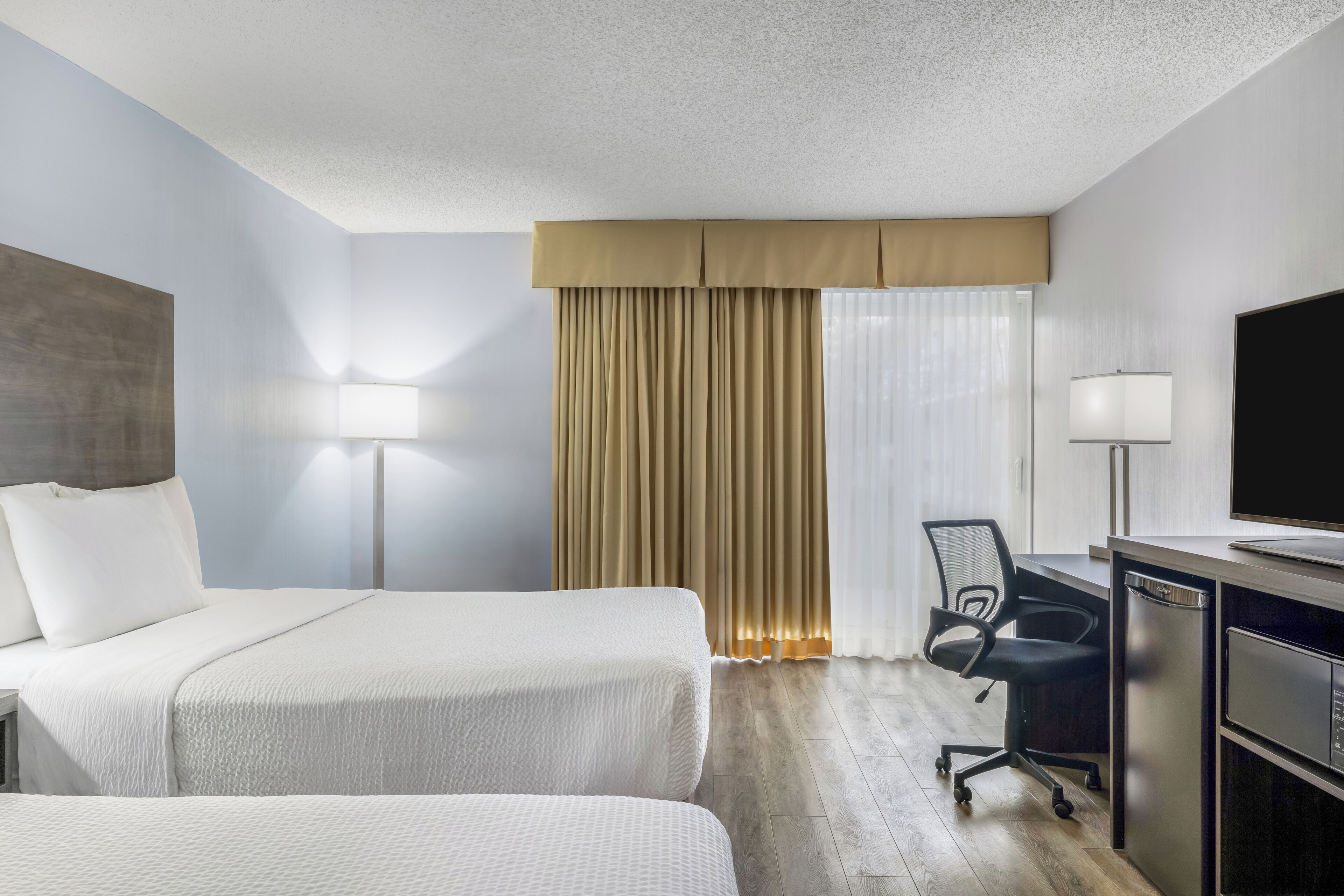 Habitación de hotel con dos camas, un escritorio con silla, televisor y ventana con cortinas.