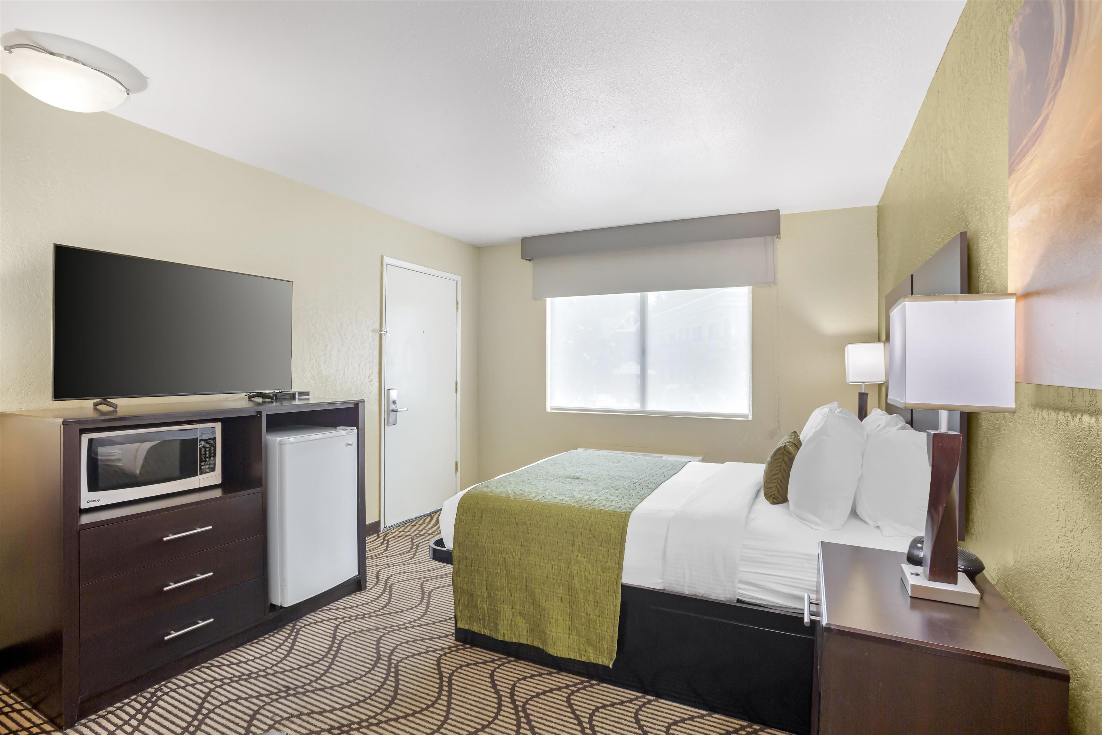 A hotel room with a bed, a flat-screen TV, a mini-fridge, a desk with a lamp, and a window with blinds.