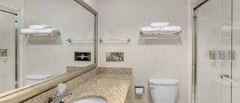 Un baño moderno con encimera de granito, un lavabo blanco, un espejo grande, un inodoro y una cabina de ducha de vidrio.