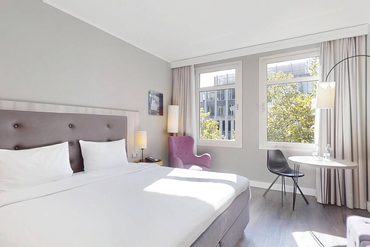 A hotel room with a large bed, a chair, a small table, and a window with a view of buildings and trees.