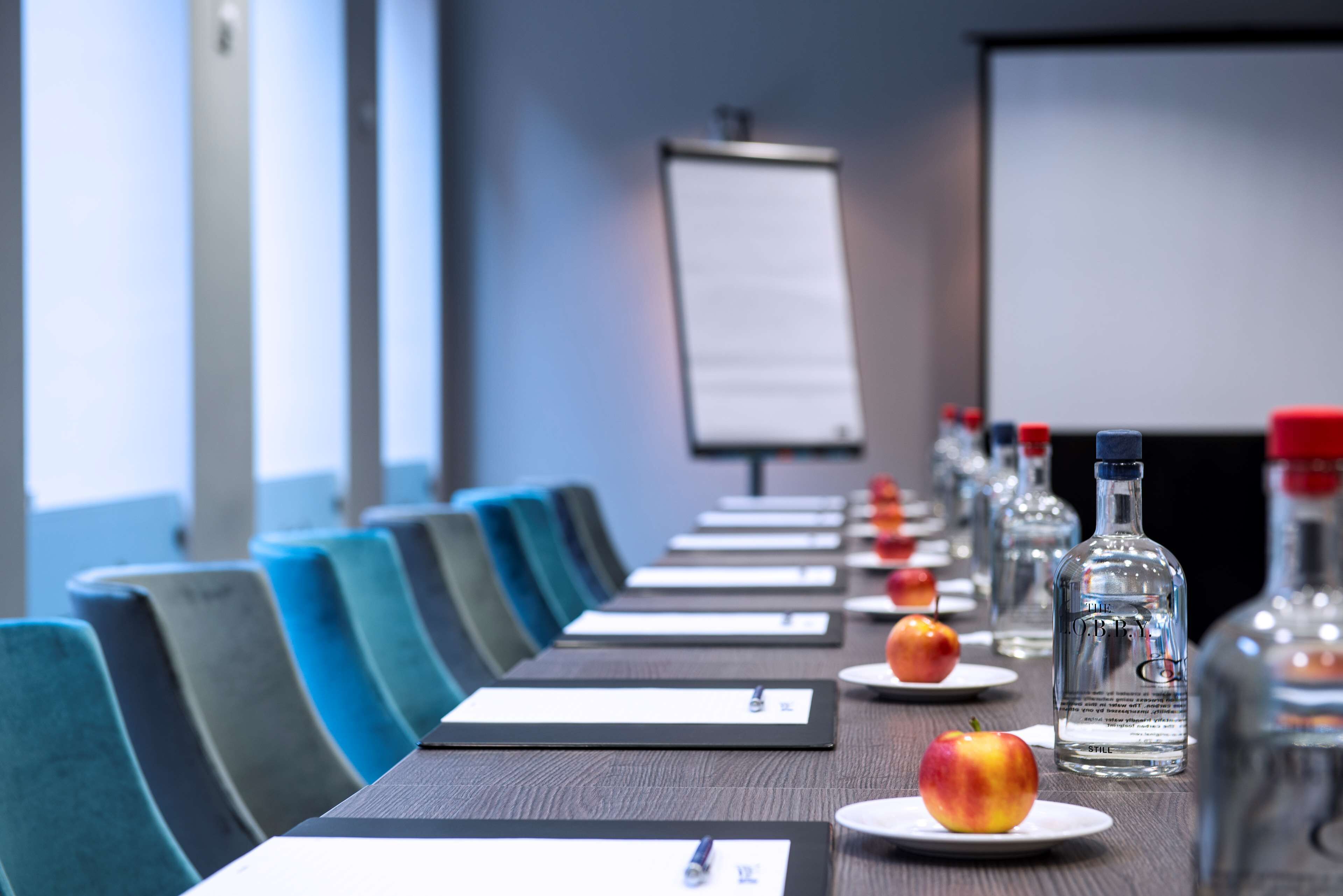 Ein Konferenzraum mit einem langen Tisch, Stühlen, einer Projektionsleinwand sowie Wasser- und Spirituosenflaschen.