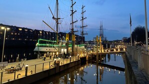 Marina frente al agua con barcos de gran tamaño amarrados, un paseo peatonal y una masa de agua tranquila que refleja el entorno.