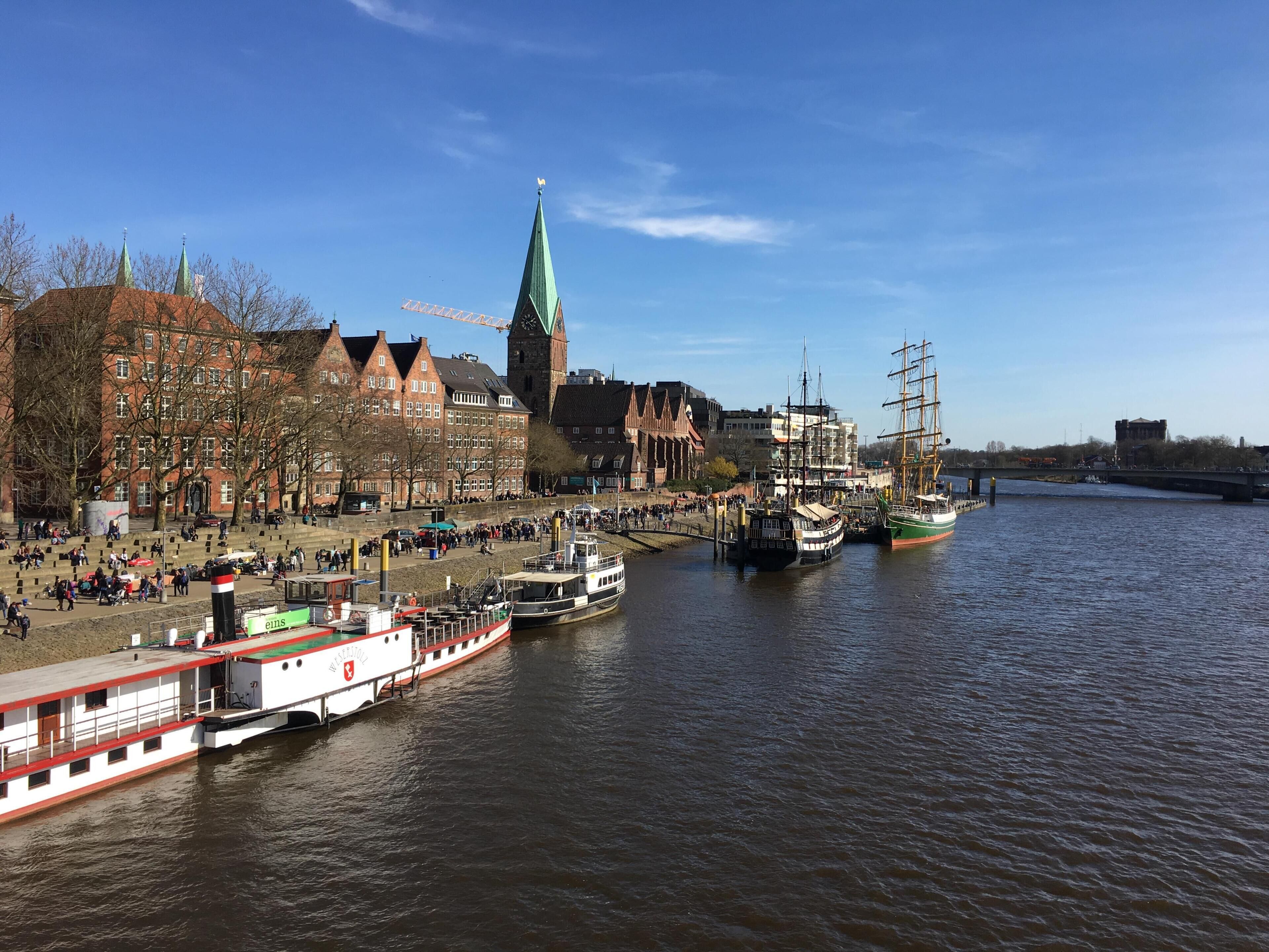 A riverside town with historic buildings, a church tower, and a river with boats.