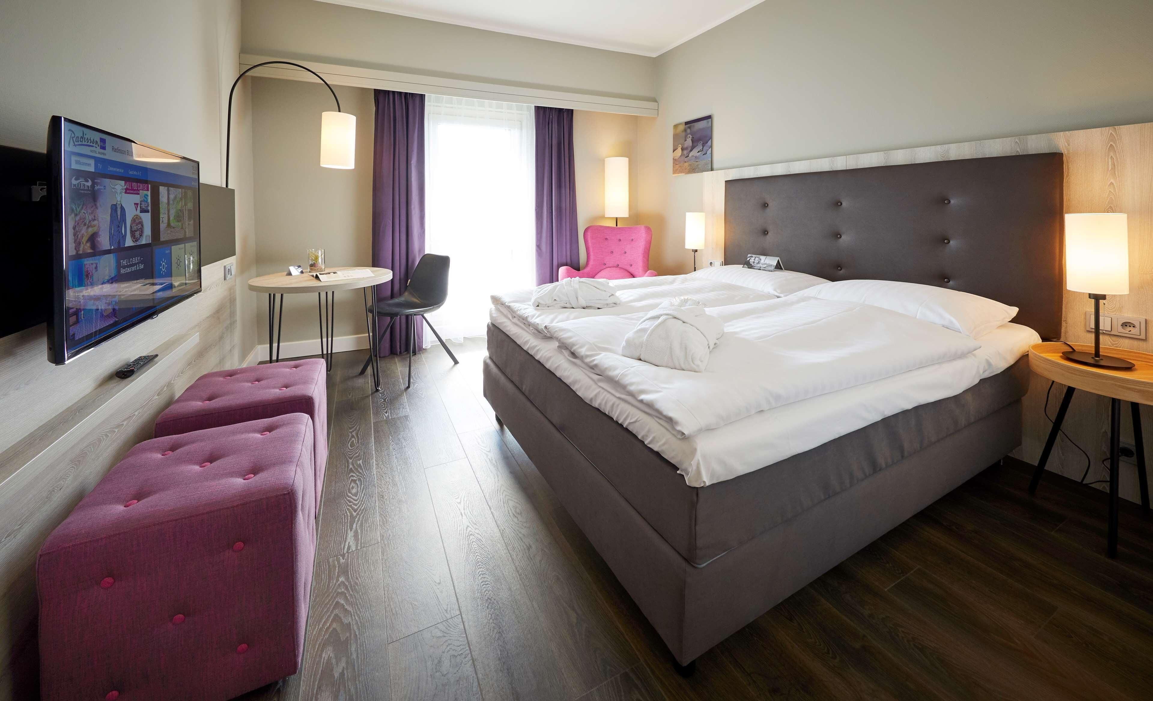Habitación de hotel con cama, escritorio, silla, televisión y ventana con cortinas.