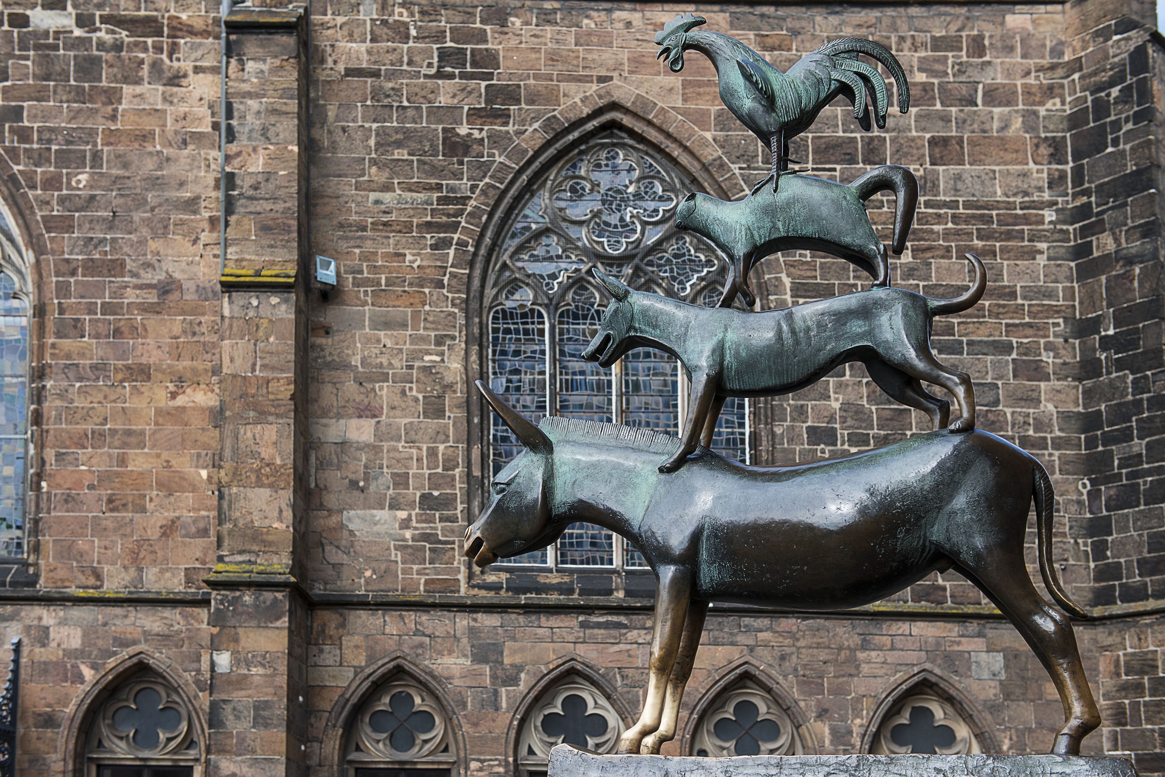 Eine bronzene Ram- und Hahnenstatue auf einem Steinsockel vor einem Backsteinhaus mit Rundbogenfenstern.