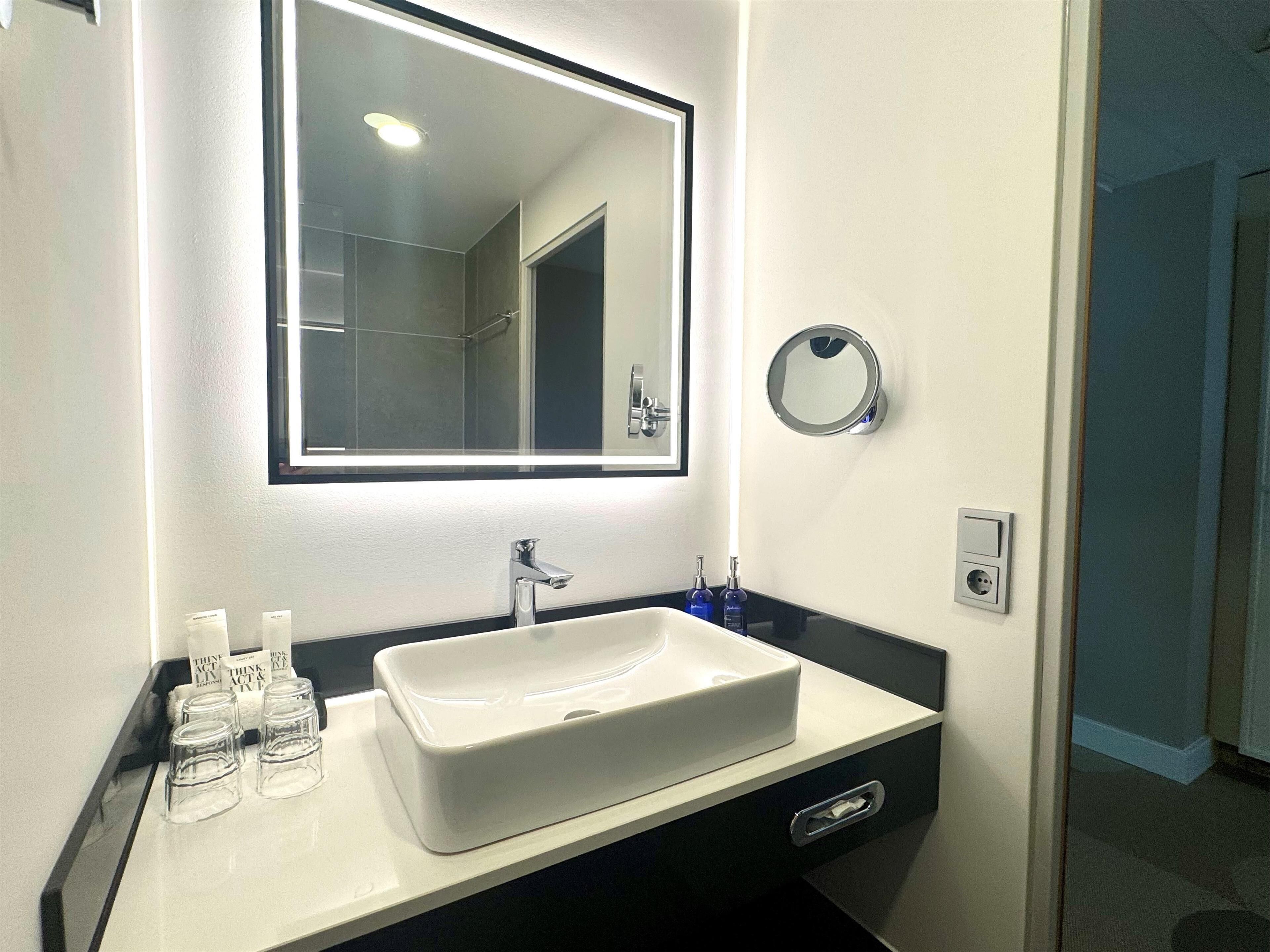 A modern bathroom with a white sink, a large mirror, and a round mirror on the wall. There are toiletries on the counter and a towel dispenser.
