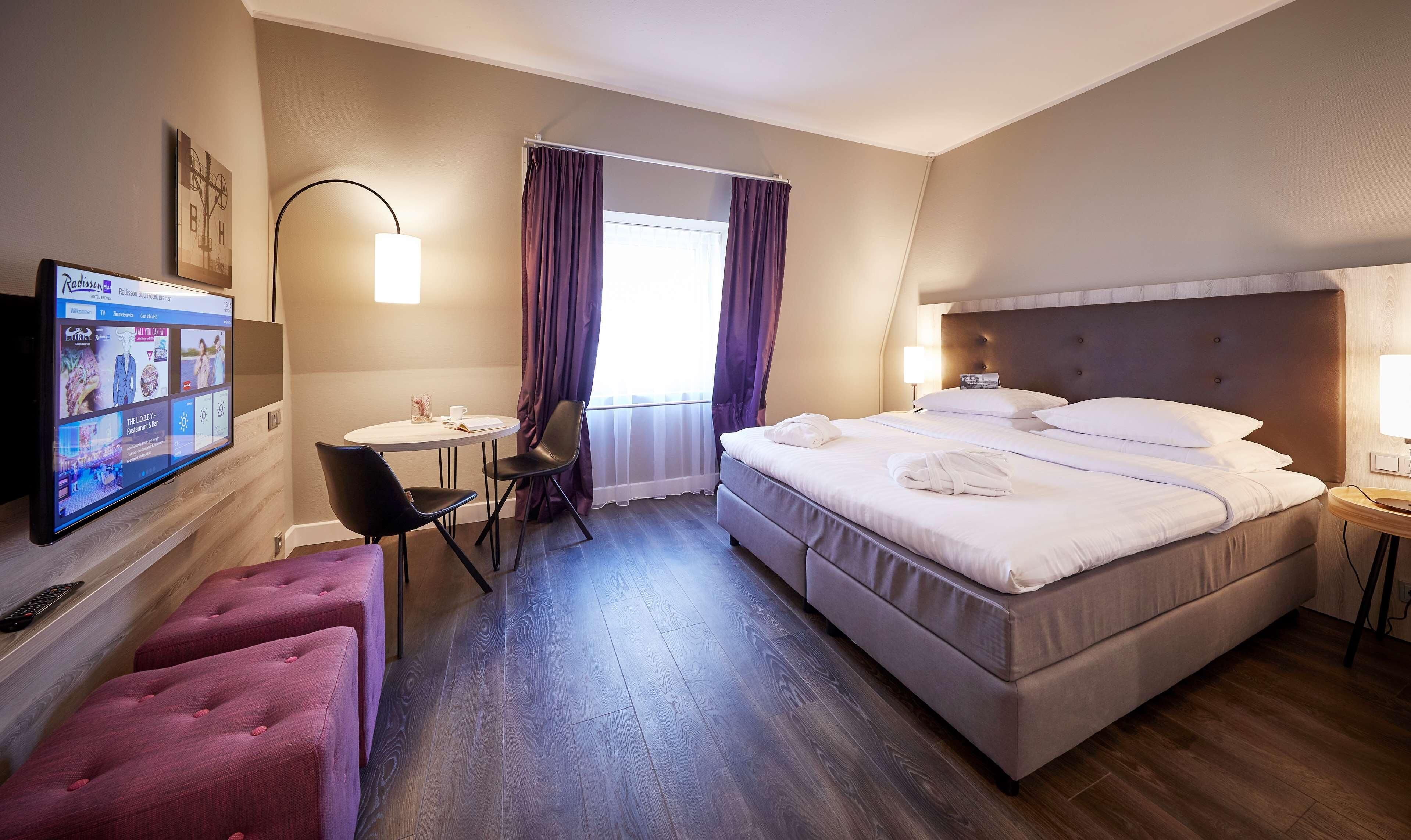 Habitación de hotel con cama, escritorio con silla, televisión y ventana con cortinas.