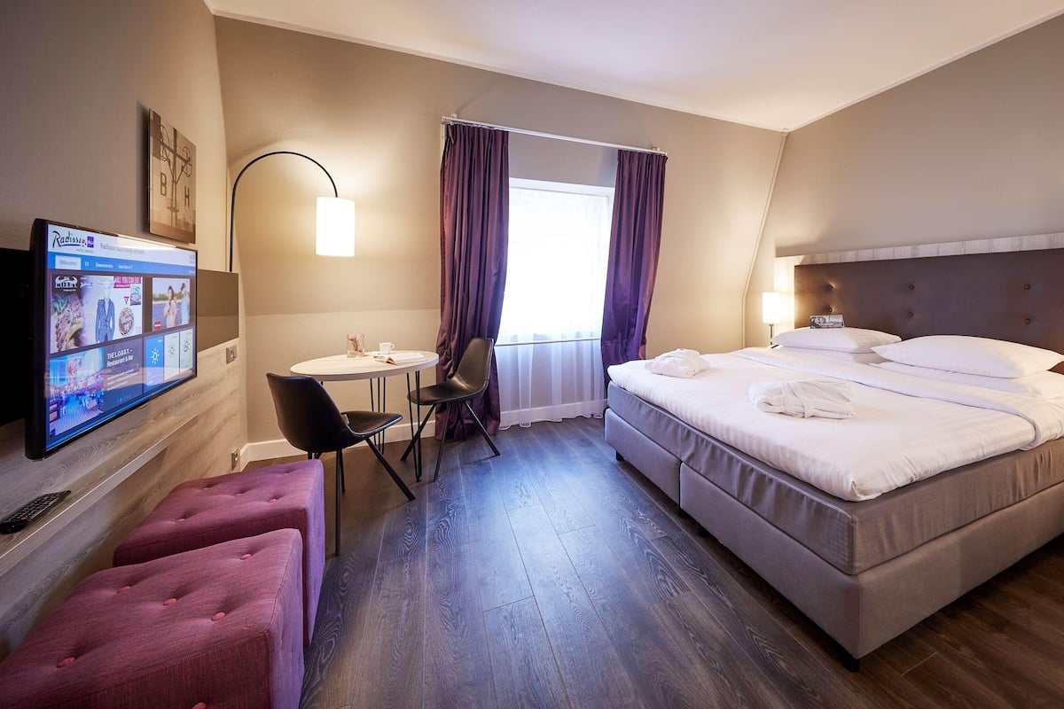 A hotel room with a bed, a desk with a chair, a TV, and a window with curtains.