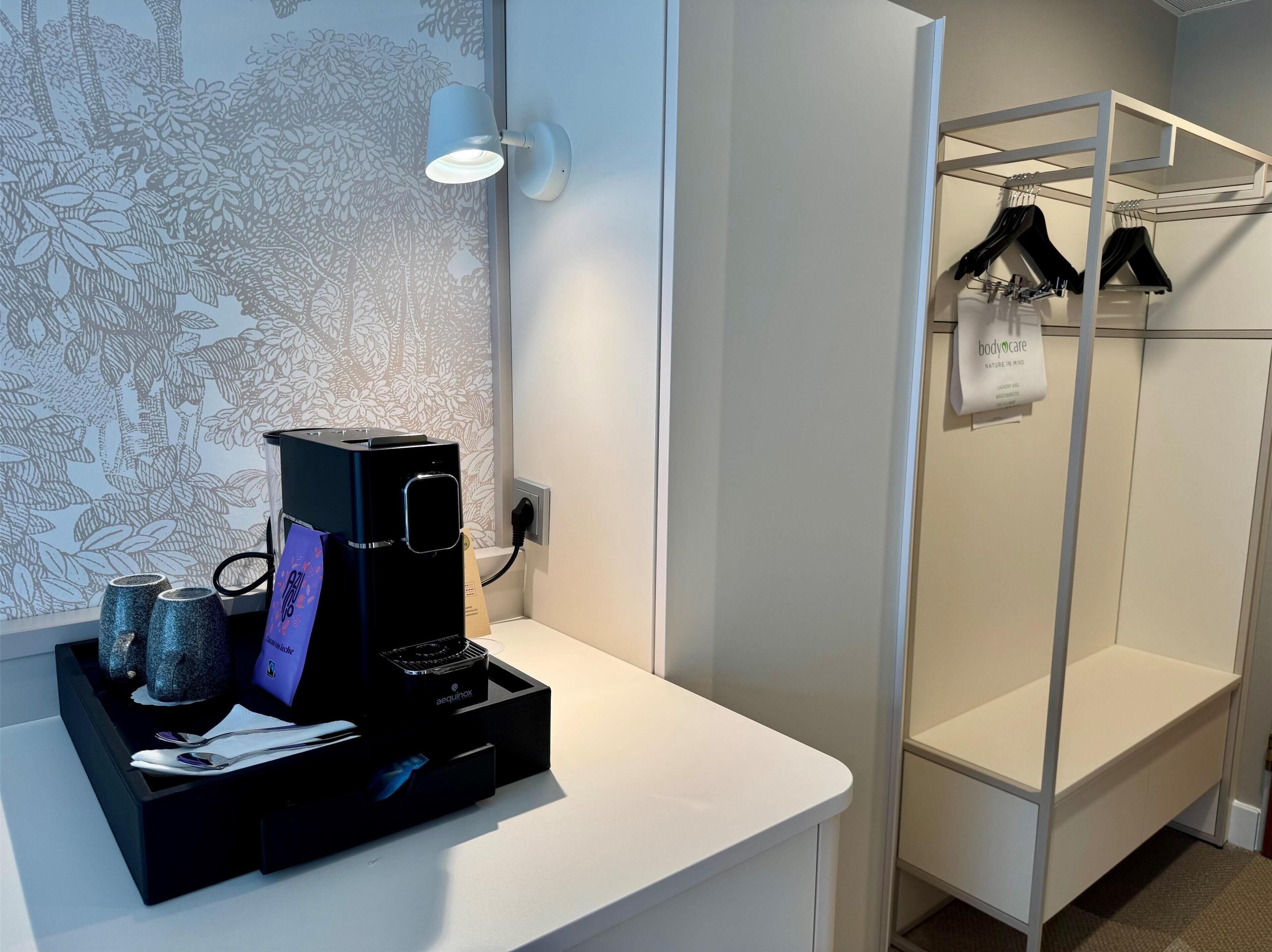 A modern bathroom with a black coffee maker on a white countertop, a towel rack with clothes, and a wall with a leaf pattern.