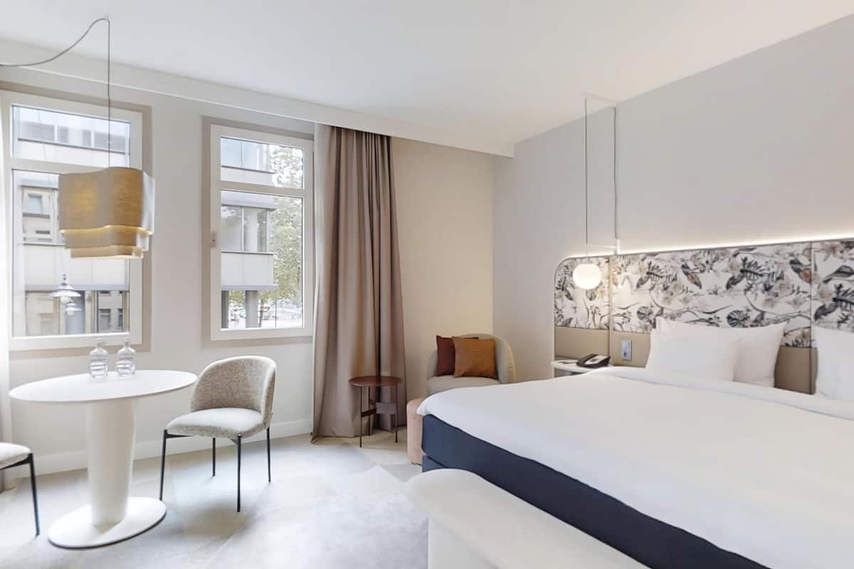 A hotel room with a large bed, a round dining table, two chairs, a small side table, and a window with curtains.