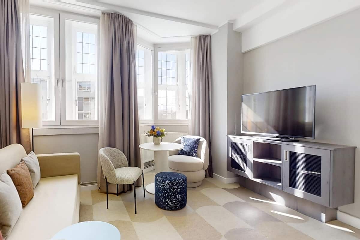 A modern living room with a sofa, a small round table with a vase, a TV on a cabinet, and large windows with curtains.