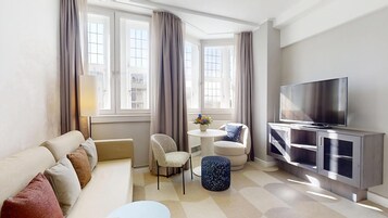 A modern living room with a sofa, a small round table with a vase, a TV on a cabinet, and large windows with curtains.