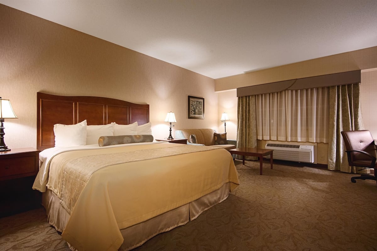 A hotel room with a large bed, two bedside tables with lamps, a desk with a chair, a window with curtains, and an air conditioning unit.