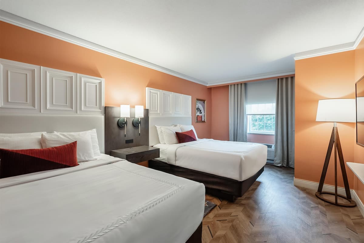 A hotel room with two beds, a wooden herringbone floor, a window with curtains, and a wall-mounted lamp.