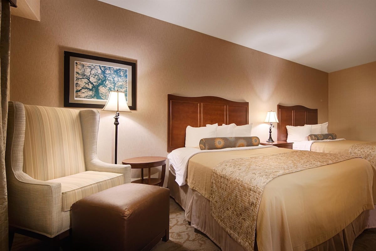 A hotel room with two beds, a chair, a small table, and a lamp.