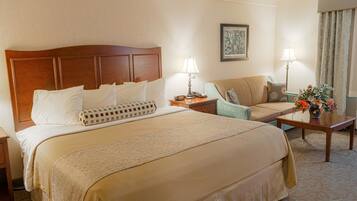 A hotel room with a large bed, a sofa, a nightstand, a lamp, and a small table with flowers.